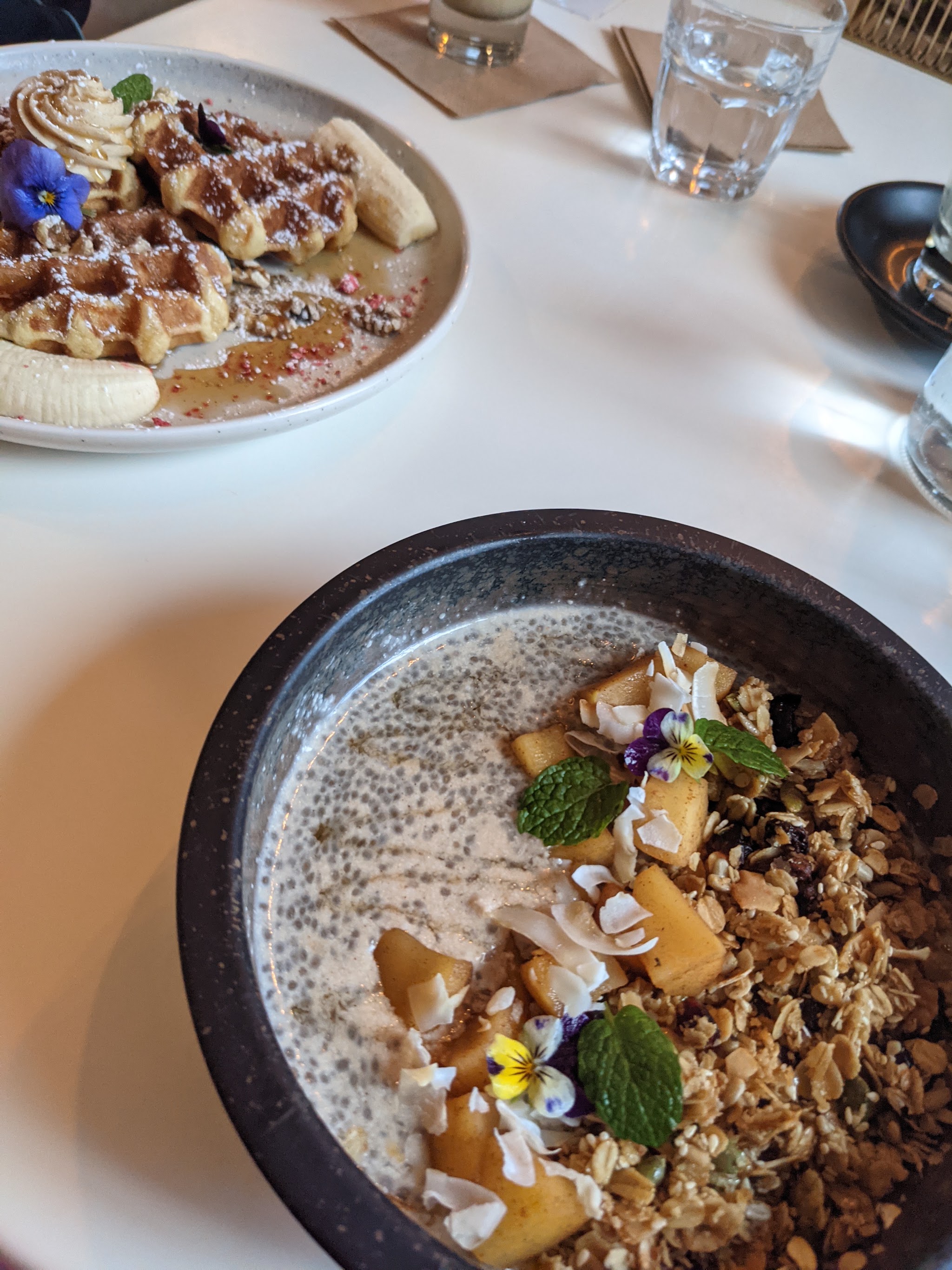 Waffles with banana and cream beside a chia bowl with granola and edible flowers